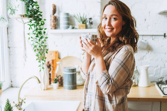 Femme souriante dans sa cuisine boit une tisane qui pourraient aider à accélérer ou à faire venir ses règles plus vite