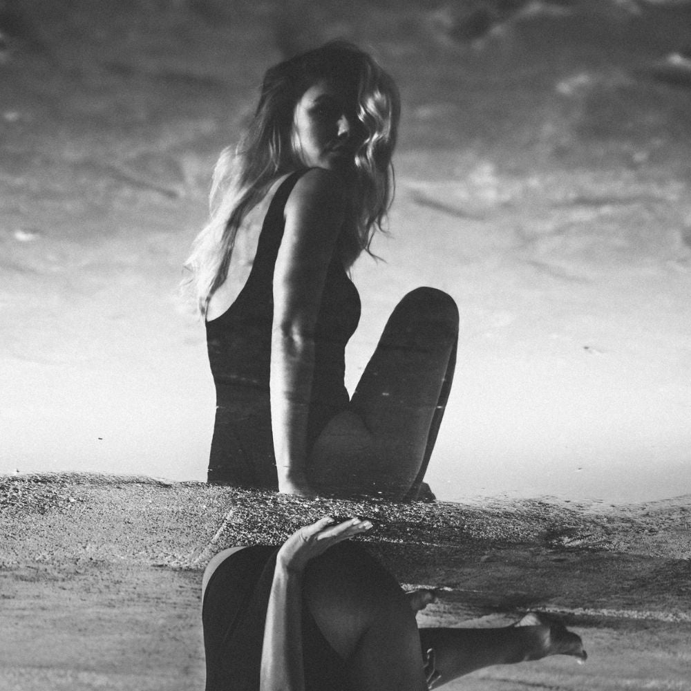 Frau im Urlaub sitzt am Strand und trägt ihren einteiligen Perioden-Bademode mit Riemchen