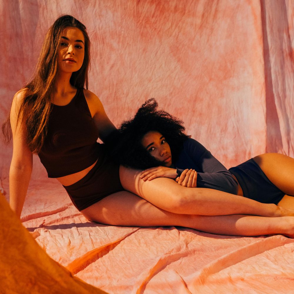 Two women posing together, one of them wearing menstrual shorts, in a warmly lit setting with a textured pink background.